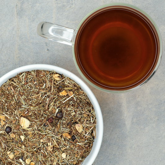 Dried cornsilk, cleavers, couchgrass, lemon verbena and juniper berries in a bowl with a cup of brewed tea on a gray surface