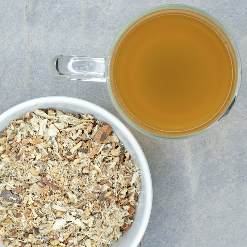 Bowl of dried wild cherry bark, marshmallow root, licorice root herbals next to a glass of lemon-colored hot tea on a gray surface
