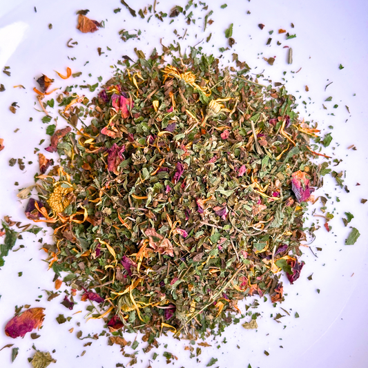 Close-up of Ease Herbal Tea in a bowl with vibrant dried rose petals, lemon balm, calendula, and peppermint leaves with a touch of purple from the gotu kola, creates a colorful and textured mosaic ofhand cut and blended herbs and blossoms.