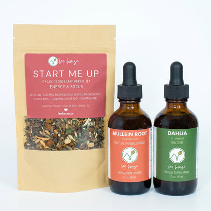A kraft paper pouch of Lee House Start Me Up herbal tea stands next to two amber glass dropper bottles with black caps. The pouch has a red label, and the bottles have orange and green labels.