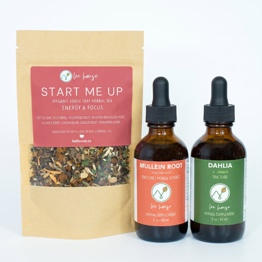 A kraft paper pouch of Lee House Start Me Up herbal tea stands next to two amber glass dropper bottles with black caps. The pouch has a red label, and the bottles have orange and green labels.
