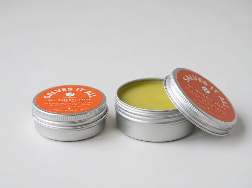 Two small round silver metal tins of Salves It All All Natural Salve on a light gray background, with one tin open to show the yellow-green salve inside and the other closed with an orange label.