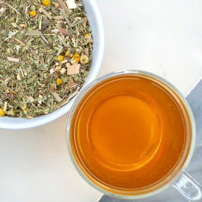 A cup of steeped golden herbal tea beside a bowl of Dusty's Dream Tea blend, highlighting the loose leaves of chamomile, linden, and other organic ingredients on a light surface.